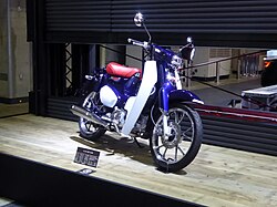 A two-tone blue and dark blue scooter with a red seat. It is shiny and well lit as if in a museum or automotive convention.