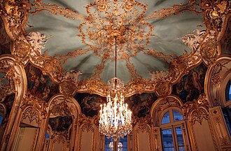 The upper white walls and gold panelling of an oval room, curving seamlessly into a domed ceiling adorned with gilded stucco vines and framed oil paintings.