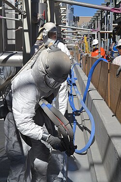 Photograph: In a narrow open passageway, two people in hazardous-material suits spray a grey substance on the floor.