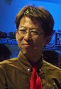 A 50-year-old Japanese man with black-brown hair in a brown suit with a red tie, smiling away from the camera.