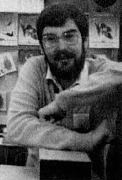 Grainy black and white photograph of a dark haired man with a short beard and glasses leaning forward on a table.