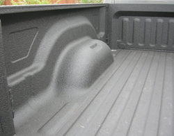 Photograph of the interior of a pickup bed covered in a gray substance. The shape of the bed is molded to allow the upper part of the wheel. There are structural ridges molded in the walls and floor.