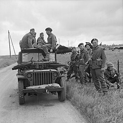 A black and white photograph of a jeep with soldiers sitting on top and standing beside it. The soldiers sitting on the jeep are three German soldiers and one British soldier who is interrogating the Germans. On the bonnet of the jeep is small motorcycle, while in the background is a Horsa glider